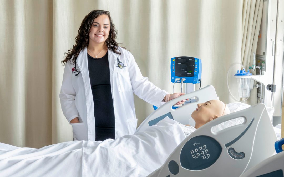 image of a student wearing a white lab coat and stethoscope standing next to a nursing simulator
