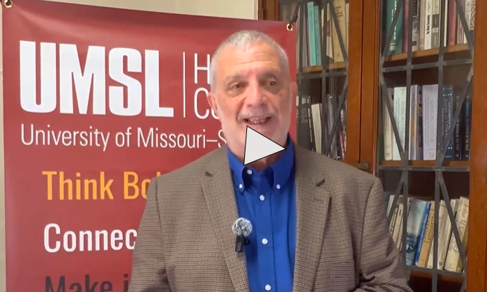 Video thumbnail showing a person standing indoors in front of a red UMSL Honors College banner and a bookshelf filled with books. The banner displays “UMSL University of Missouri–St. Louis” and Honors College branding. A play button icon is centered over the image, indicating a video.