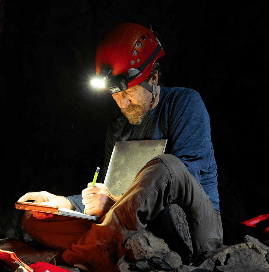 A cave explorer in a red helmet uses a headlamp to take notes while sitting on rocky terrain.