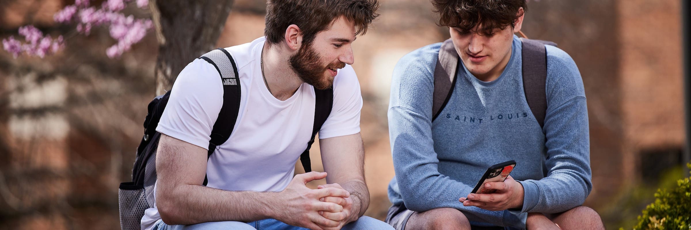 Outside on a bench, a student shows another student his phone
