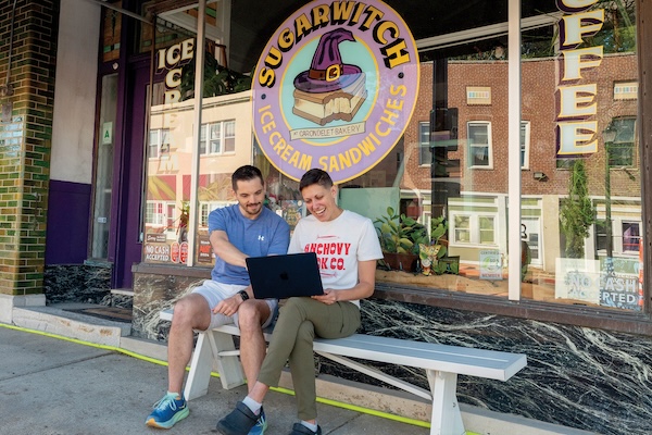 Two people sit on a bench outside Sugarwitch, an ice cream sandwich shop, while looking at a laptop together.