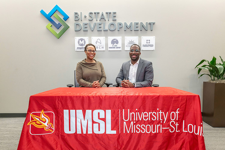 Two people seated behind a table with a red UMSL (University of Missouri–St. Louis) tablecloth at Bi-State Development.