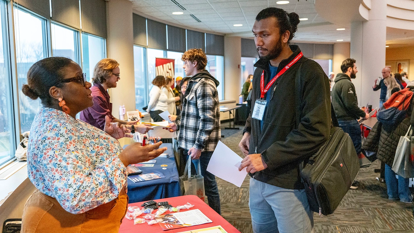 Orientation fair scene with a woman discussing opportunities with a male attendee, surrounded by various informational displays and other participants interacting in the background.