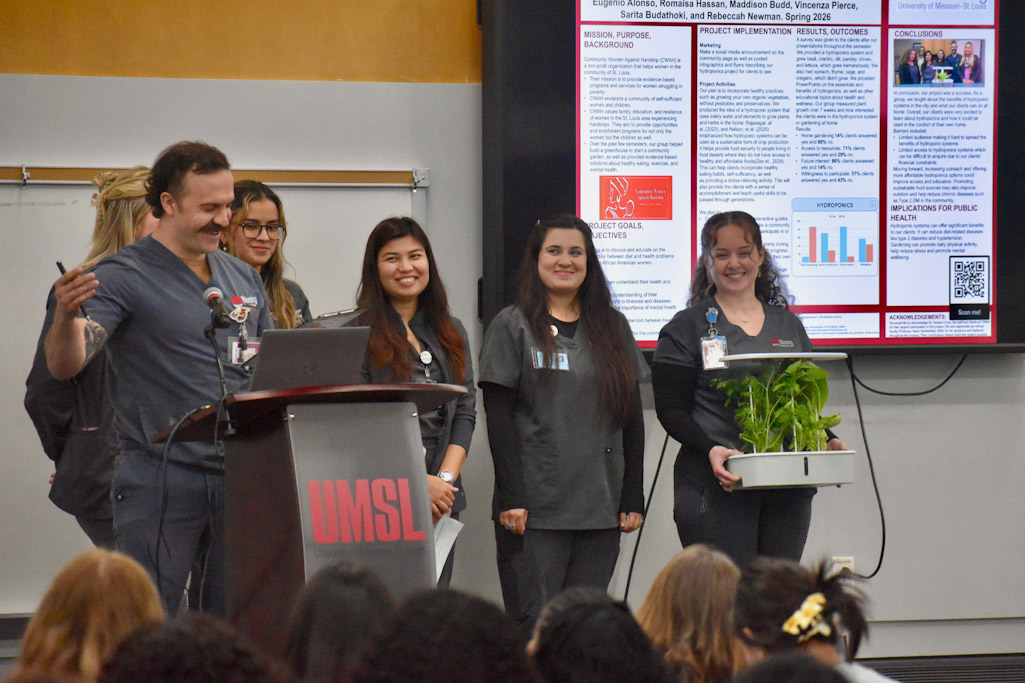 A group of presenters stands on a stage at UMSL, with one speaking at a podium and another holding a small tray of leafy plants, while a research poster is displayed on a screen behind them.