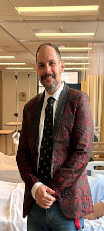 Man in a patterned burgundy blazer and tie standing beside a hospital bed in a clinical room with privacy curtains and medical poles.