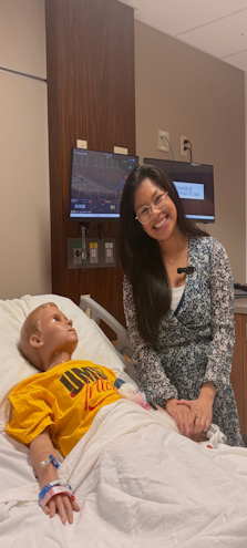 Smiling woman in a patterned dress stands beside a hospital bed holding the hand of a medical training mannequin, with patient monitors visible in the background.