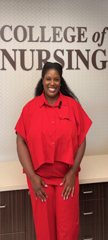 Smiling woman in a red outfit standing in front of a wall sign that reads “College of Nursing.”