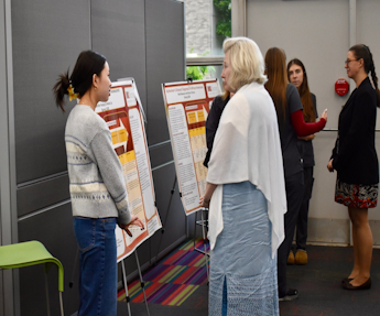 People stand and talk near research posters on easels in a hallway, as a presenter faces one poster while others gather nearby.