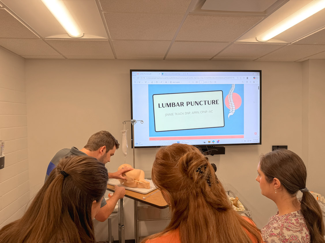 Healthcare trainees watch an instructor demonstrate a lumbar puncture on a mannequin in a classroom, with a slide titled "Lumbar Puncture" displayed on a wall monitor.