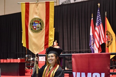 Graduate in cap and gown smiling at a commencement ceremony, with university banners and U.S. flags on stage behind her.