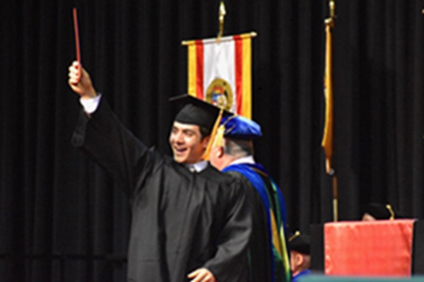 Graduate in cap and gown on a stage raising a diploma cover during a commencement ceremony.