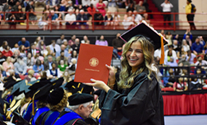 Graduate in cap and gown smiles while holding up a diploma cover during a commencement ceremony in a crowded indoor arena.
