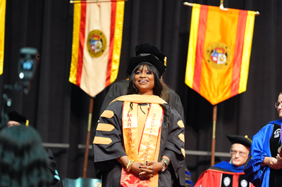 Graduate in a black doctoral gown and cap stands smiling on a stage, wearing an orange stole, with ceremonial banners hanging behind them.