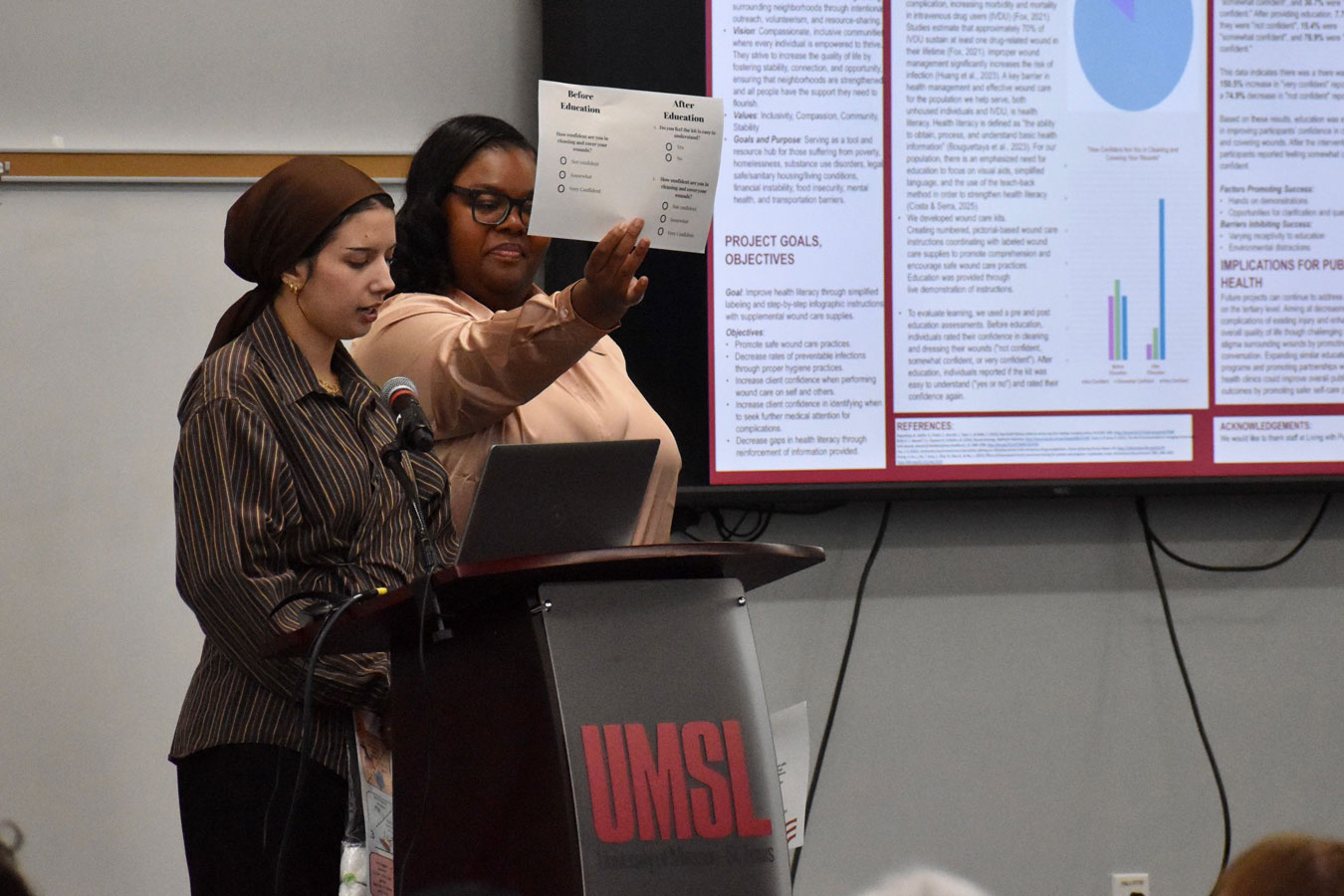 Senior-Nursing-Students-Destini-and-Nesreen-Presenting-Their-Project-ting toward a large screen displaying charts and text.