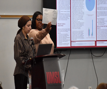 Two women give a presentation at a podium labeled UMSL, with one pointing toward a large screen displaying charts and text.