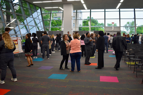 People mingle and chat at a networking event inside a modern lobby with large floor-to-ceiling windows and colorful carpet tiles.