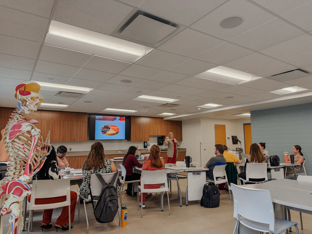 Classroom scene with students seated at tables while an instructor presents near a screen showing a pie chart; an anatomical skeleton model stands in the foreground.