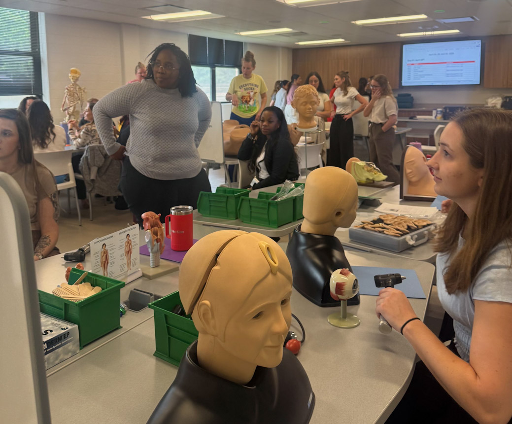 Students in a classroom lab gathered around tables with medical training mannequins and anatomy models, including a head and an eyeball.