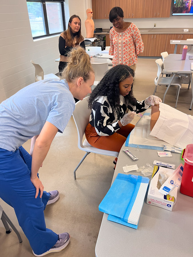 Students in a classroom lab practice a medical procedure on a mannequin arm at a table with gloves and supplies while instructors observe.