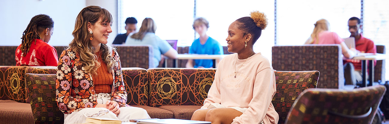 2 students sitting on a couch chatting, other students in the background