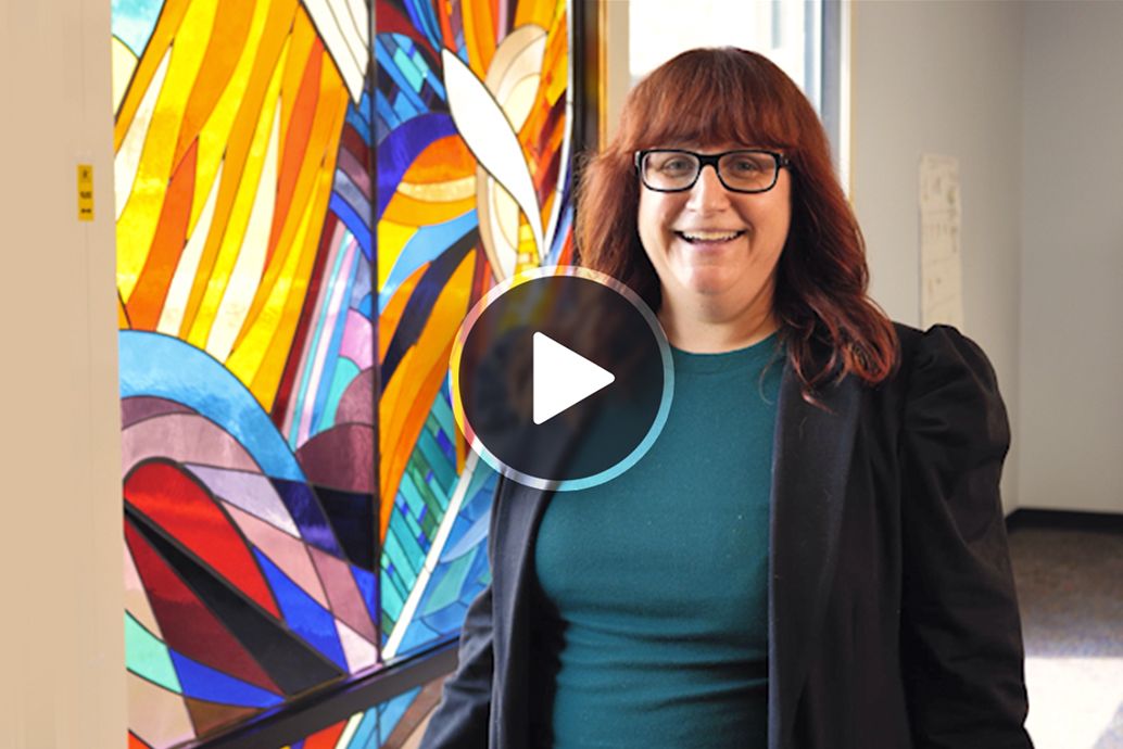 Amber candela smiling with glasses standing indoors beside a colorful stained-glass wall, with a play button overlay indicating a video thumbnail.