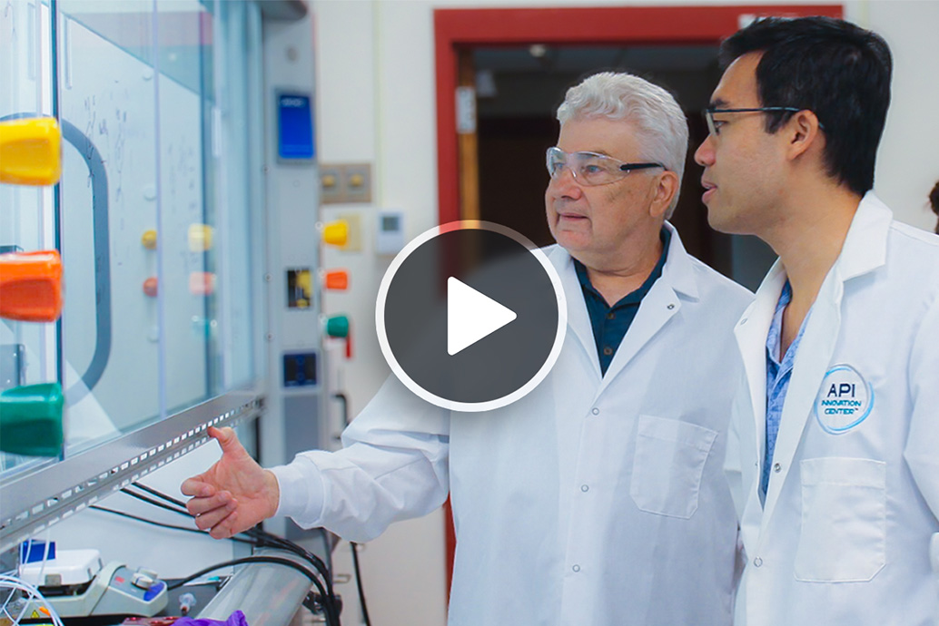 Two scientists in lab coats review equipment behind a glass safety panel in a laboratory.