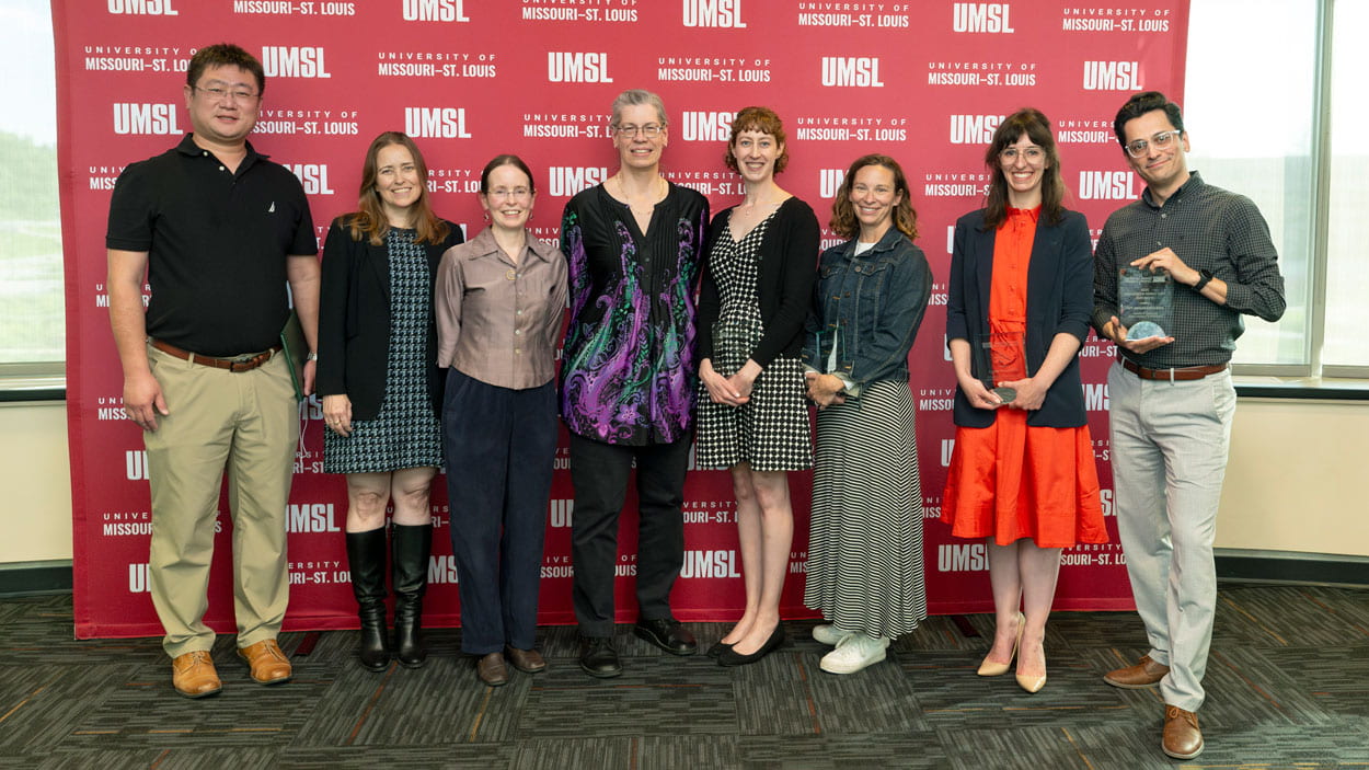 Group of eight people posing at an awards event in front of a University of Missouri–St. Louis (UMSL) step-and-repeat backdrop, with one person holding a glass trophy.