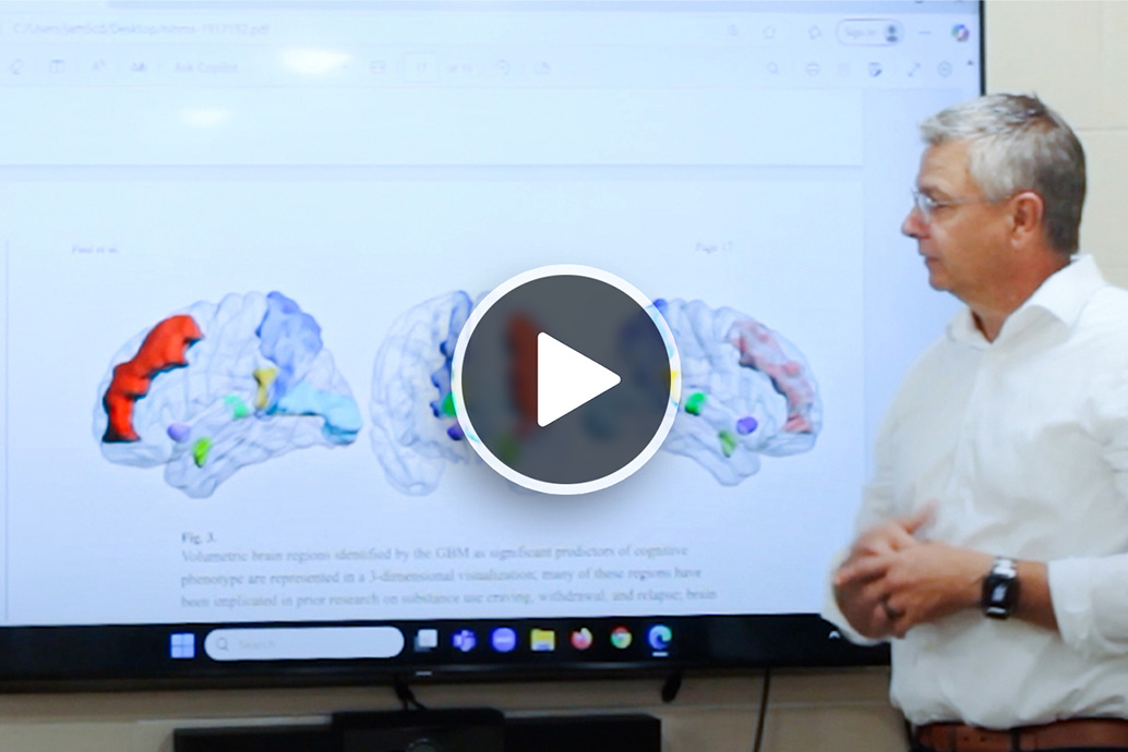 Researcher giving a presentation next to a large screen showing three brain visualizations with highlighted activity regions.