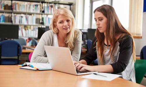 Two women seated at a table near a library bookshelf. The younger woman types on her laptop while the other woman looks on.