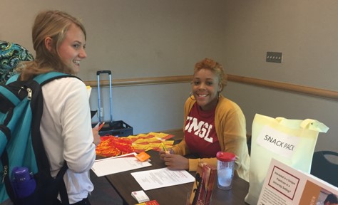 A student wearing a UMSL shirt smiles while interacting with another student at a registration table with promotional materials and snack packs.