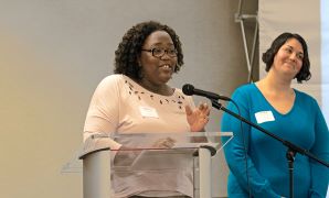 Two women are speaking at a podium during a presentation, with one actively engaging the audience and the other smiling in support.