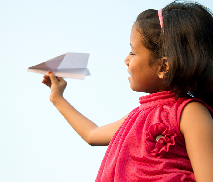 Young child in a pink top holds a paper airplane out in front of her against a clear sky.