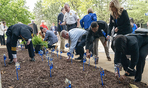 Group of people and uniformed officials place small blue pinwheels into a mulched garden bed along a walkway.