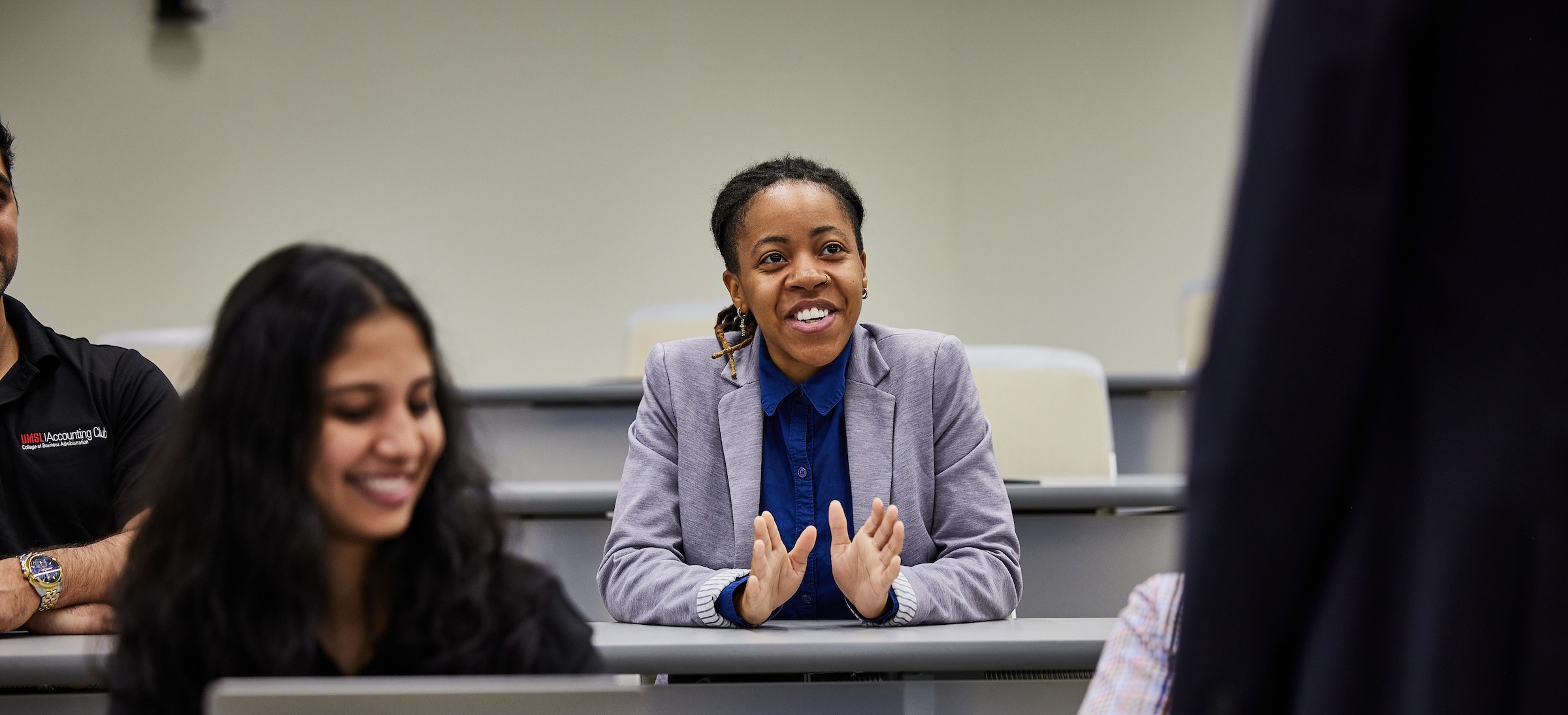 A student in a gray blazer sits in a classroom, smiling and speaking while gesturing with their hands. Other students sit nearby, including one in the foreground using a laptop and another partially visible to the side. The atmosphere appears engaged and conversational during a class discussion.