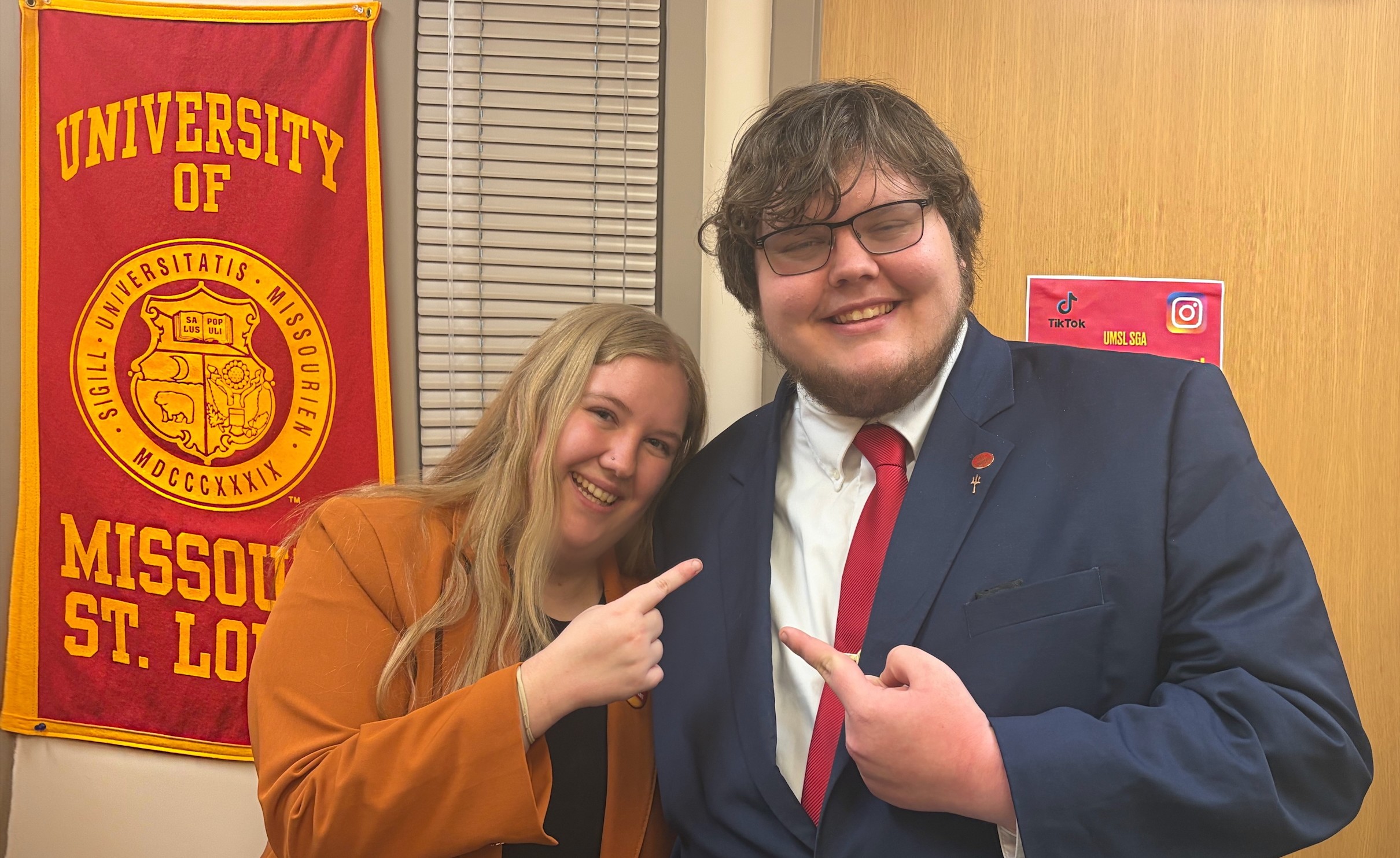 Two students stand side by side in a hallway, smiling and pointing at each other in a playful, celebratory pose. The student on the left wears an orange blazer and has long blond hair. The student on the right wears a navy suit with a red tie and glasses. Behind them hangs a red and gold University of Missouri–St. Louis banner, and a small sign with TikTok and Instagram logos is posted on the door.