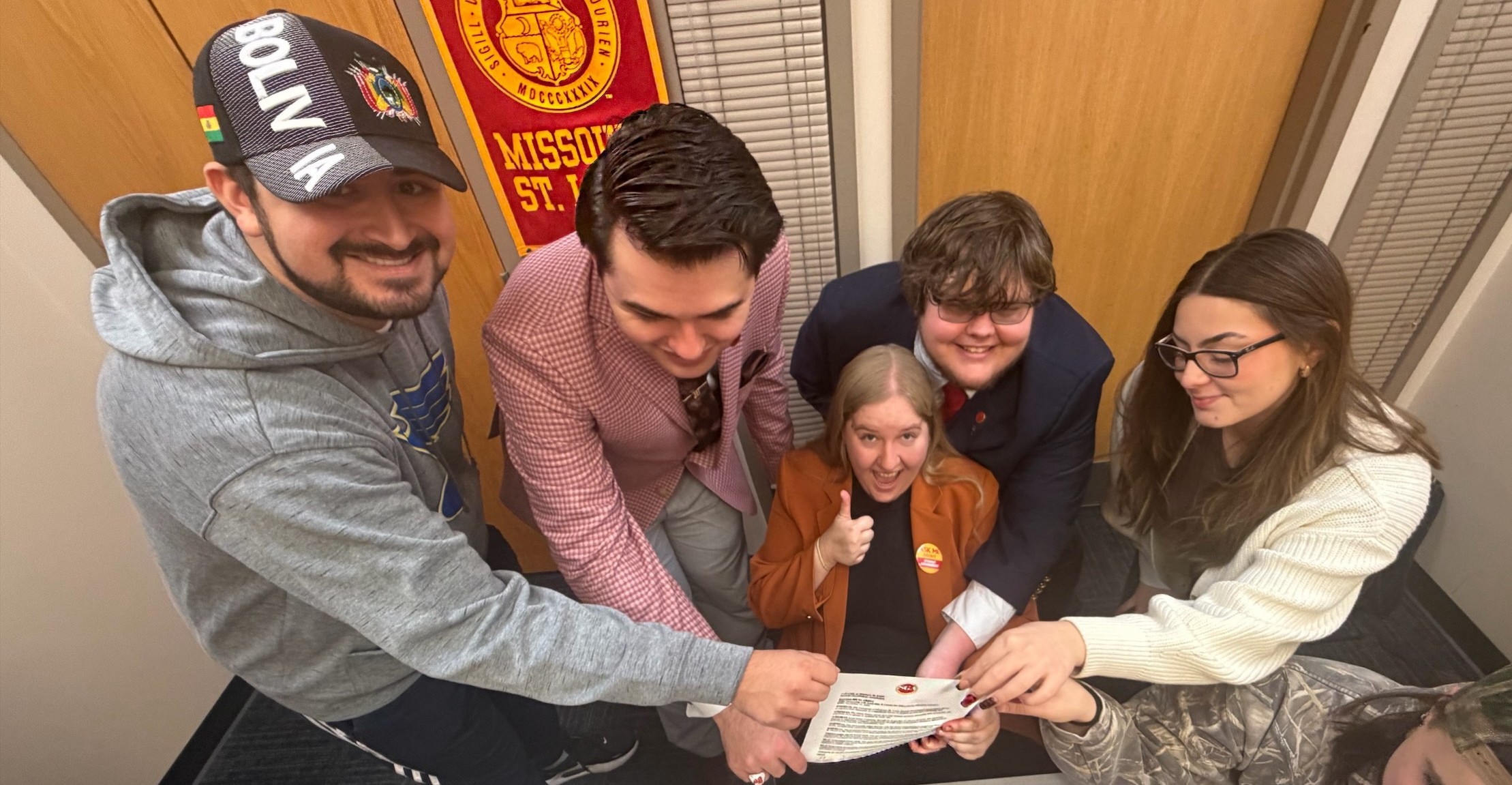 A group of six students gather closely in a hallway, smiling and looking down at a document they are holding together. One student wearing a “Bolivia” hat and a gray hoodie reaches in from the left, while others dressed in business or casual attire lean in from different angles. A student in an orange blazer gives a thumbs-up toward the camera. A Missouri St. Louis banner hangs on the wall behind them. The group appears excited and celebratory.