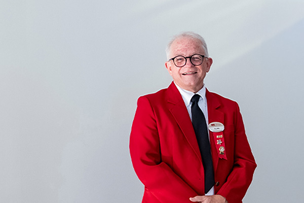 Smiling older man in a red blazer and tie standing against a plain light background.