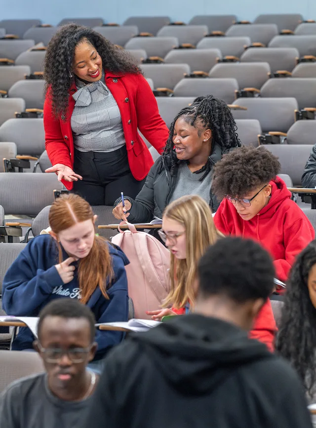 Instructor in a red blazer talks with students as they take notes in a lecture hall.