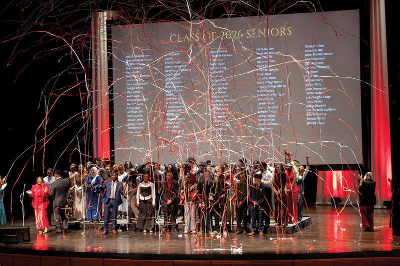 Graduating seniors on a stage as red and white streamers fall in front of a screen reading “Class of 2026 Seniors.”