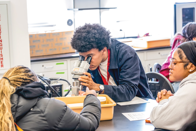 Student looks through a microscope during a science lab while classmates watch at the table.