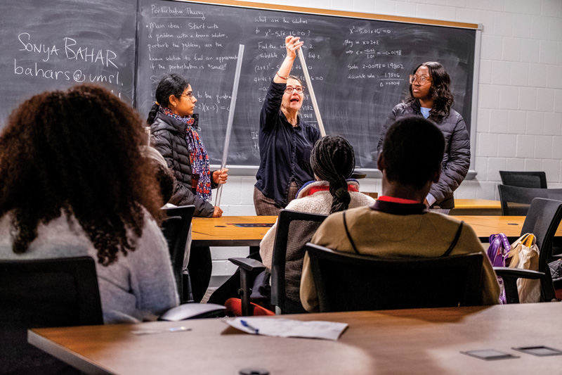 Instructor demonstrates a math concept with measuring tapes in front of a chalkboard while students watch in a classroom.