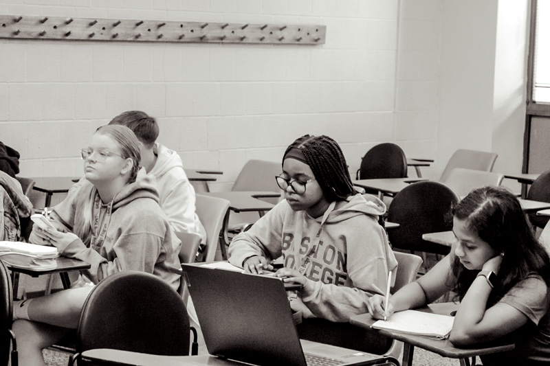 Students in a classroom taking notes and working on laptops at individual desks.