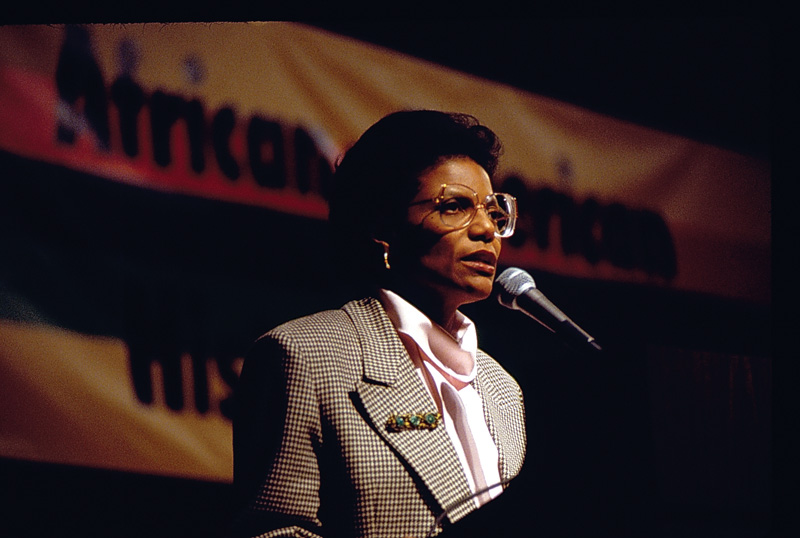 Woman in a checkered blazer speaking into a microphone onstage in front of a blurred banner.