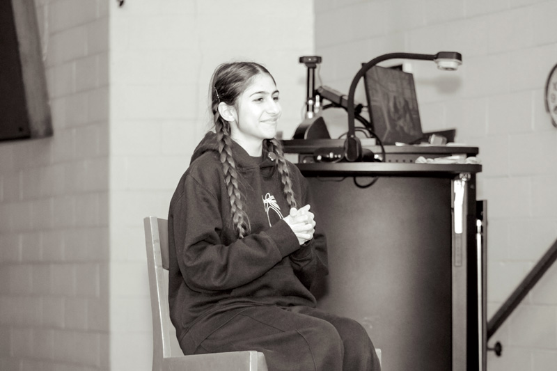 Teen girl with braided hair sitting and smiling near a classroom lectern with a computer and microphone.