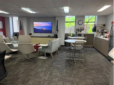 Lounge-style office break room with sofas facing a wall-mounted TV, café tables and chairs, and a kitchenette along the right wall.