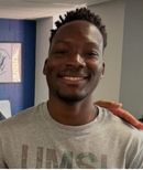 Smiling person in a gray T-shirt indoors, with a hand resting on their shoulder.