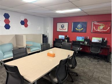Office meeting room with a large table, black chairs, a lounge nook, and a row of computer workstations against a red accent wall with military-themed flags.