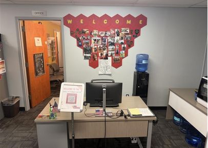 Office reception desk with a computer monitor, water cooler, and a wall display of photos spelling “WELCOME” behind it.