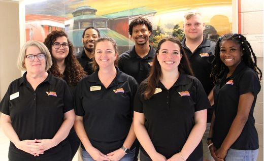 Group of eight staff members in matching black polo shirts posing and smiling indoors, with a colorful mural in the background.
