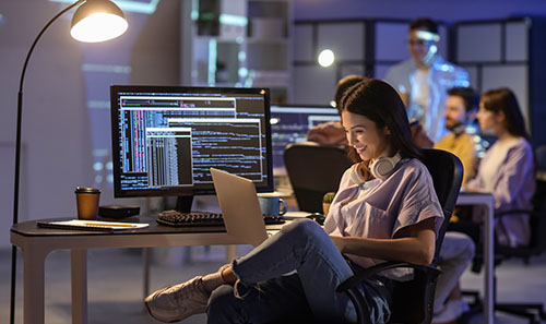 A young techy type female sits at her desk typing code on her laptop.